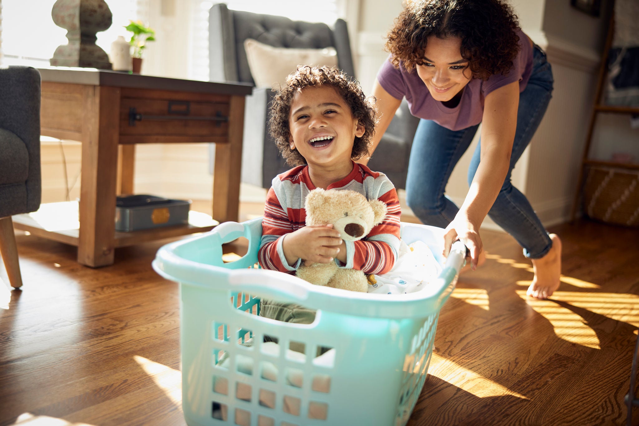 Older sister gives younger brother a ride in the laundry basket. African American teenage girl gives her younger brother a ride in a laundry basket. She pushes him around in a laundry basket with his stuffed bear on the hardwood floor of the home where they live.
