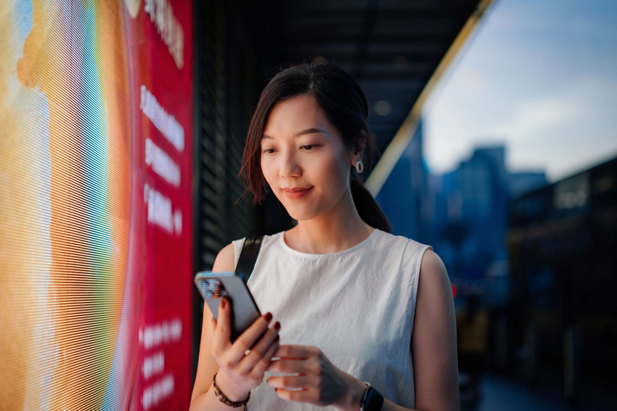 Confident young Asian businesswoman using smartphone in downtown city street in the evening, standing against illuminated LED digital display in the city. Lifestyle and technology