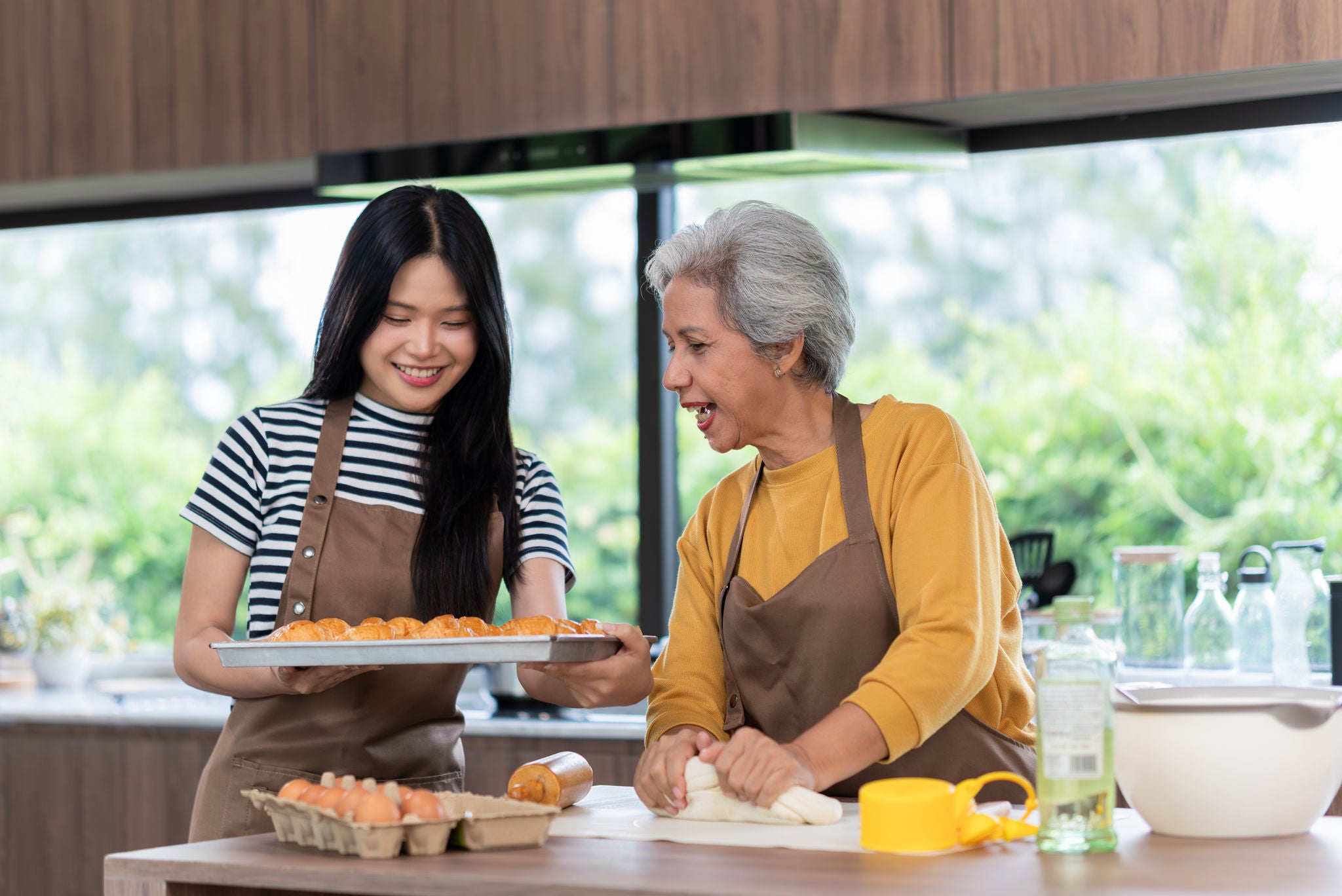 A cheerful moment between an elderly Asian woman and her granddaughter as they bake pastries together in a modern home kitchen. Wearing aprons and surrounded by baking ingredients, the two share laughter and connection through a fun, everyday activity. The warm light and casual atmosphere highlight the joy of intergenerational bonding, family support, and shared traditions.