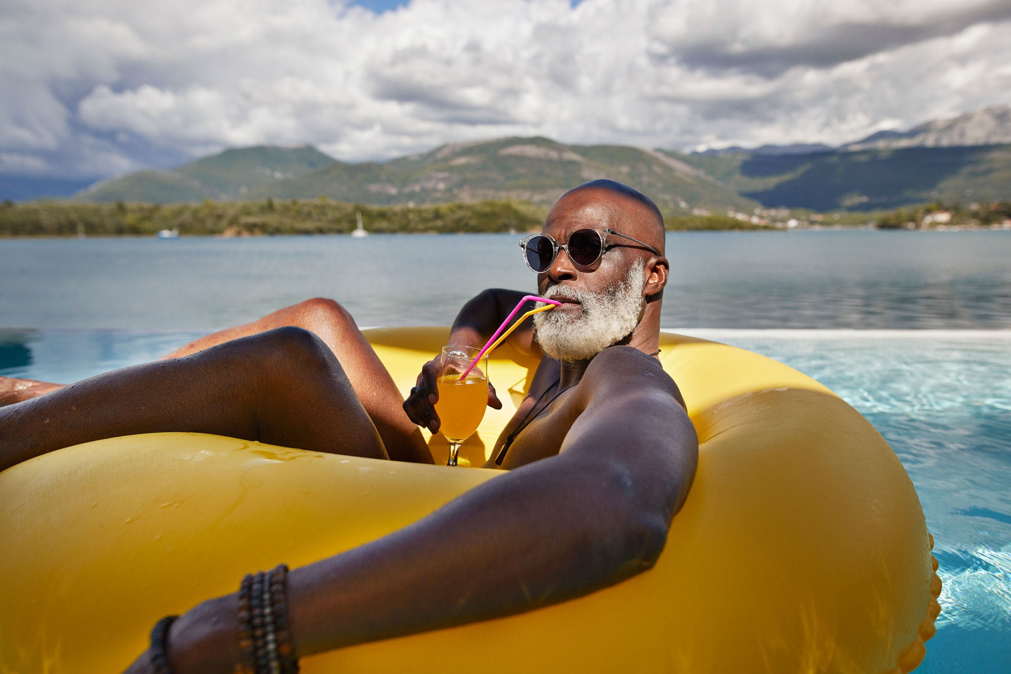 Bald senior man wearing sunglasses while drinking juice in inflatable ring during vacation