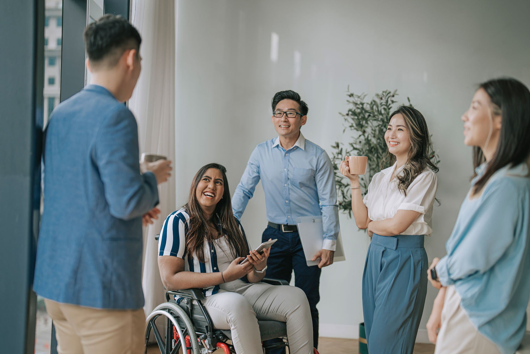 Indian white collar female worker in wheelchair having cheerful discussion leading conversation with colleague in creative office workstation beside window