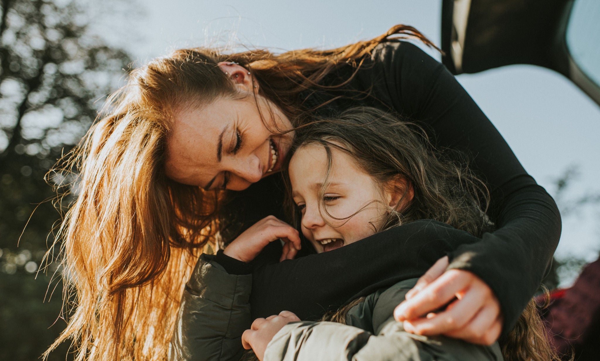 A young Mum bends over to her daughters height, and wraps her arms around her. They both wear thick winter coats as they brace the cold weather, and warm up in a loving embrace.