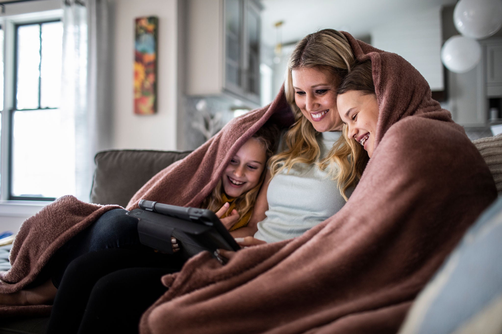 Mother and tween daughters snuggled under blanket and watching a movie on digital tablet